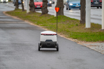 An autonomous modern food delivery robot moves along the sidewalk in rainy weather. Concept of...