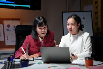 Two businesswomen working together using laptop in office meeting room at night, discussing new project strategy and taking notes