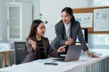 Two young Asian businesswomen collaborating and discussing projects while working together on a laptop in a modern office setting