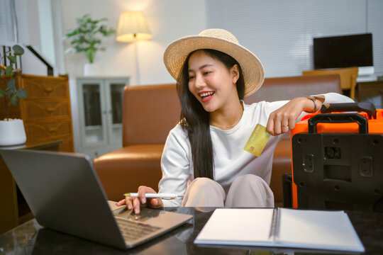 Happy tourist planning a vacation while using a laptop and holding a gold credit card beside a suitcase, filled with excitement and anticipation