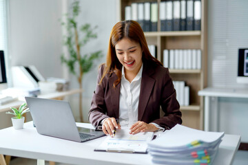 Young businesswoman smiling and reviewing financial documents and working on a laptop at her desk in a modern office