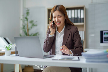 Young happy manager smiling while talking on mobile phone and using laptop computer in modern office