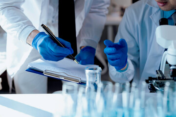 Two scientists wearing lab coats and gloves are analyzing data and taking notes on a clipboard in a...