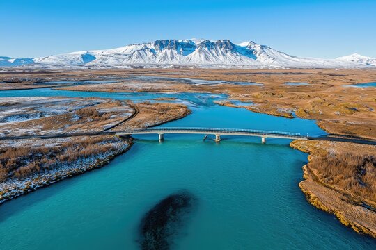 Aerial view of a bridge spanning a vibrant river amidst snowy mountains.