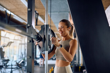 Fototapeta premium Happy sportswoman adjusting weight disk on barbell during strength training in gym.