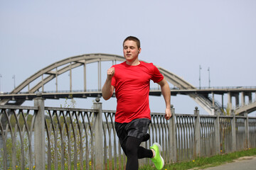 Young sportsman running near river and bridge, wearing red t-shirt. The guy is doing a running...