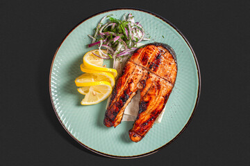 A perfectly grilled salmon steak served on a mint green ceramic plate, accompanied by lemon wedges and a garnish of finely sliced red onion and fresh dill. 