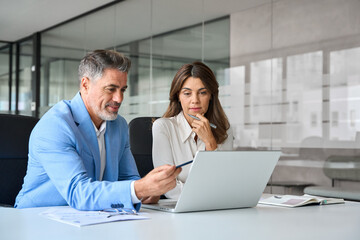 Bank managers consulting at meeting. Two busy mature professional business man and woman executives team using laptop working on computer having conversation on financial project in office.
