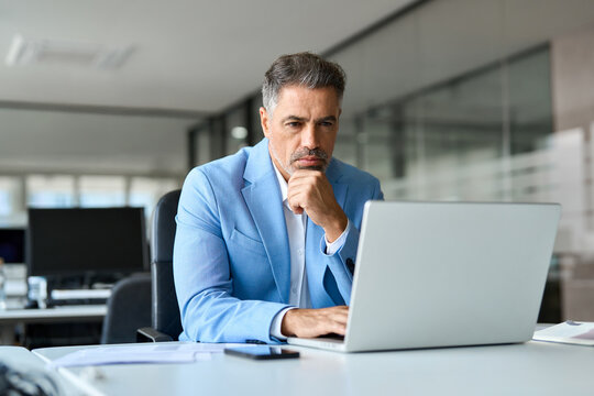 Busy 50 year old senior businessman executive, serious middle aged professional business man financial investor wearing suit using laptop computer working on investment project sitting at office desk.