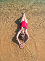 A woman in a red swimsuit lies on her back on sandy beach, enjoying the sun and warm water.