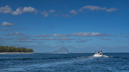 Obraz premium A tourist motorboat is sailing on the blue ocean. Foam trail behind the stern. Ripples on the surface of the water. Green vegetation on the coast of the island. Mountain against azure sky and clouds.