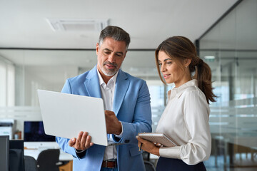 Plakat Busy professional smiling businessman and businesswoman leaders having conversation at work. Executives team of two business people working talking using laptop technology standing in corporate office