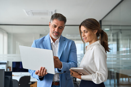Busy professional smiling businessman and businesswoman leaders having conversation at work. Executives team of two business people working talking using laptop technology standing in corporate office