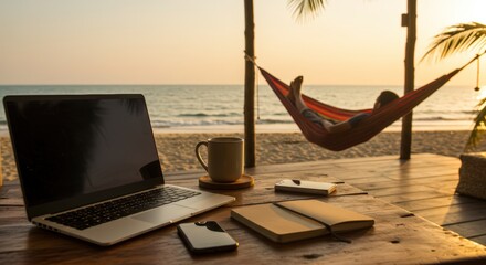 Laptop, coffee mug, and smartphone on a wooden desk with a man relaxing in a hammock by the beach at sunset. Remote work concept.