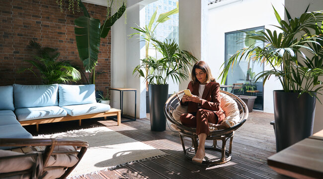 Middle aged 45 year old business woman, busy elegant mature lady wearing suit using mobile cell phone looking at smartphone sitting in comfortable chair in modern sunny office space with green plants.
