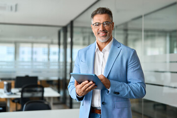 Fototapeta na wymiar Happy professional businessman ceo, older corporate leader executive manager in his 50s looking at camera. Confident mature business man investor wearing suit standing in office holding tab, portrait.