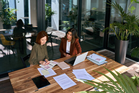 Two diverse busy business women of young and middle age talking in creative green office sitting at desk. Professional ladies employee and manager having conversation using laptop at work. Top view.