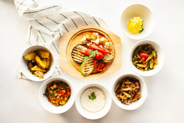 Assorted side dishes arranged in white bowls placed in a semicircle around a wooden round board with grilled vegetables. 