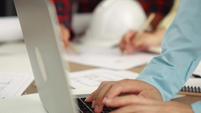 Architect and safety engineer discussion about building blueprint displaying on laptop on table with architectural document and wooden block scatter around at modern office. Closeup. Delineation.