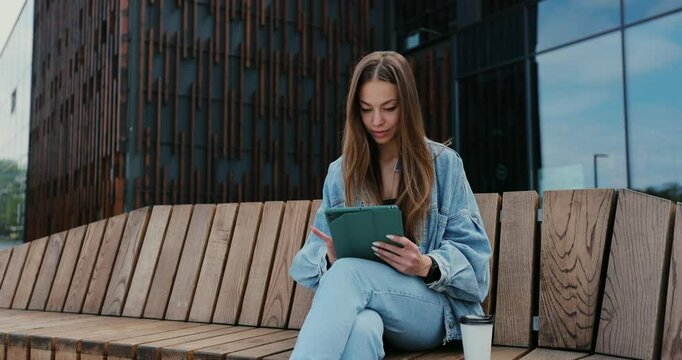 Young girl using tablet computer outdoors in the street while sitting on the bench. Pretty female tourist strolling and looking at screen, browsing Internet, chatting.