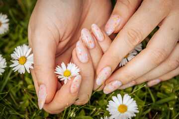 Close-up of hands with pastel pink nails featuring colorful polka dots, gently holding a white daisy on green grass, symbolizing spring beauty.
