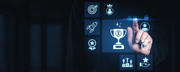 Businessman touching glowing digital award trophy icon with futuristic achievement and success symbols on dark blue interface background concept.