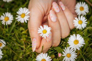 Close-up of hands with pastel pink nails featuring colorful polka dots, gently holding a white daisy on green grass, symbolizing spring beauty.