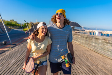 Happy skaters couple walking and laughing on a boardwalk by the sea
