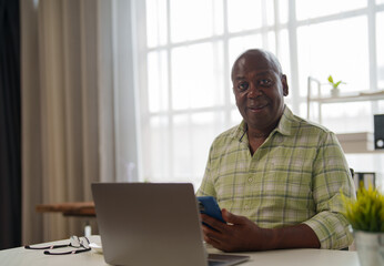 Happy african american manager sitting at desk and using laptop and smartphone, working from home, bright modern home office interior with window in background