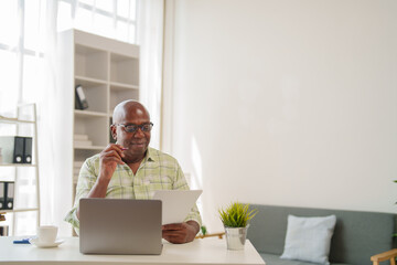 Mature African American entrepreneur reading and analyzing documents while working on a laptop at home, thoughtfully planning project strategies and reviewing financial reports
