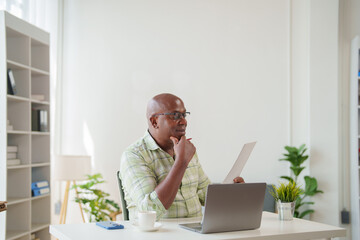 Confident mature African American businessman reading documents and working on a laptop at a modern office table, analyzing financial reports and planning project strategies
