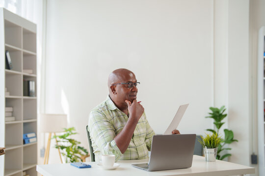 Confident mature African American businessman reading documents and working on a laptop at a modern office table, analyzing financial reports and planning project strategies