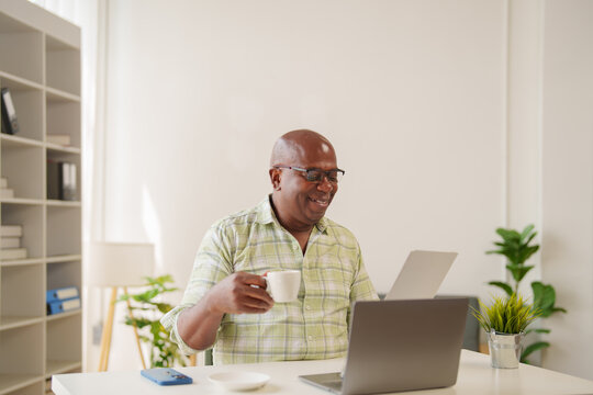 Smiling african american businessman is drinking coffee while working with laptop and documents at home office, sitting at white desk with smartphone and potted plant - Powered by Adobe