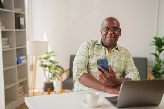 Senior black man using smartphone and laptop while sitting at desk in home office, smiling and looking at camera, with bookshelf, sofa and plant in background