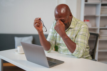 Senior black man taking off glasses and rubbing his eyes, suffering from eye strain, dry eye syndrome or headache after long laptop use at home office