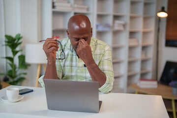 Tired senior man suffering from headache, taking off glasses and rubbing eyes after using laptop at home office, feeling eye strain, bad blurry vision, health problem concept