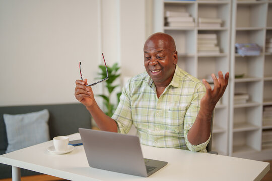 Smiling mature african american entrepreneur having a business video call on laptop and holding eyeglasses in hand while sitting at desk in home office