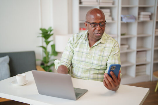 Focused mature African American man sitting at a desk, using a laptop and checking a smartphone while working remotely from a comfortable home office environment