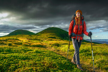 Female hiker with backpack and trekking poles walking on mountain trail under dramatic sky, wearing red jacket and beanie.