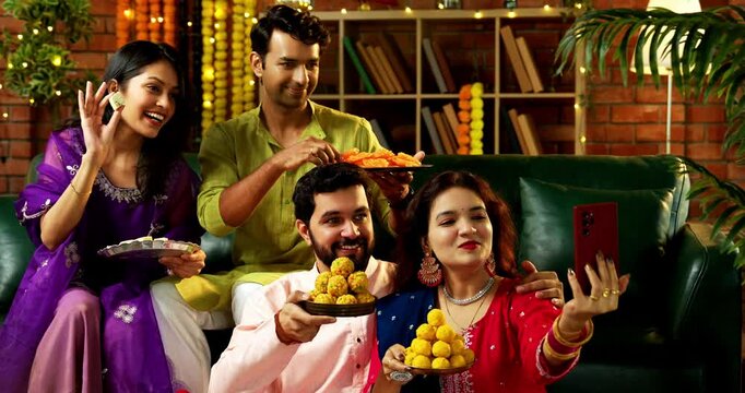 Taking Diwali selfie with friends, Indian Asian young married couple in traditional attire celebrating Hindu festival Deepavali at decorated home holding laddu barfi sweets clicking selfie