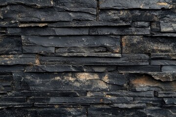 A close-up of a black slate wall with unevenly sized stones arranged in horizontal rows. The stones have a rough, textured surface and are slightly weathered, giving the wall a rustic appearance