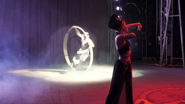 Gymnastics on wheels performed by a young man on the circus stage, a girl dancing in the foreground, a circus performance with smoke and colorful spotlights.A circus performer rotates on a cyr wheel.