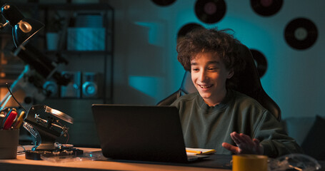 A boy chats with a friend via laptop camera, waving in greeting. They are engaged in a friendly conversation about their mutual interest in robotics and inventions.