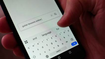 Close-up of a male hand entering the query GUITAR LESSONS ONLINE into a search bar of a browser on a smartphone