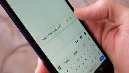 Close-up of a male hand entering the query HOW TO LOSE WEIGHT FAST into a search bar of a browser on a smartphone