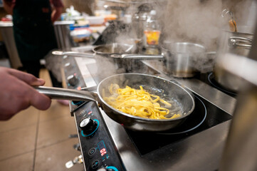 A professional chef prepares steaming pasta in a frying pan on a stovetop. The background features...