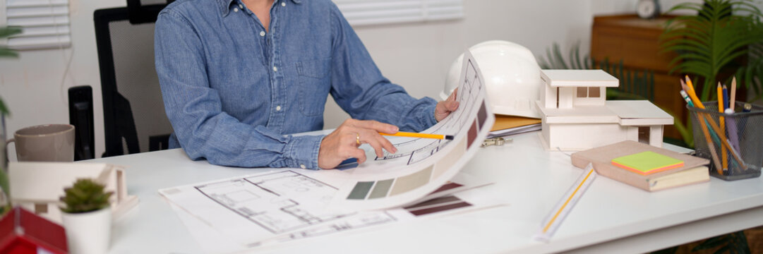 Asian Adult Architect Analyzing Blueprint for House Construction Project Design with Pencil and House Model on Desk in Office for House Interior Design - Powered by Adobe