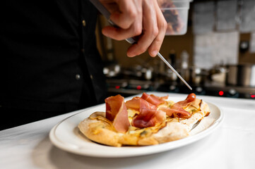 A professional chef carefully prepares a dish, adding slices of cured meat to a flatbread in a...