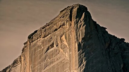 Granite cliff face of a towering mountain with rugged textures and dramatic shadows during golden hour, under a pale sky