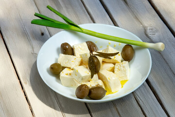 A bowl contains cheese, olives, and green garlic. The white brine cheese and olives were poured over with extra virgin olive oil, and green garlic lay on the edge of the bowl.